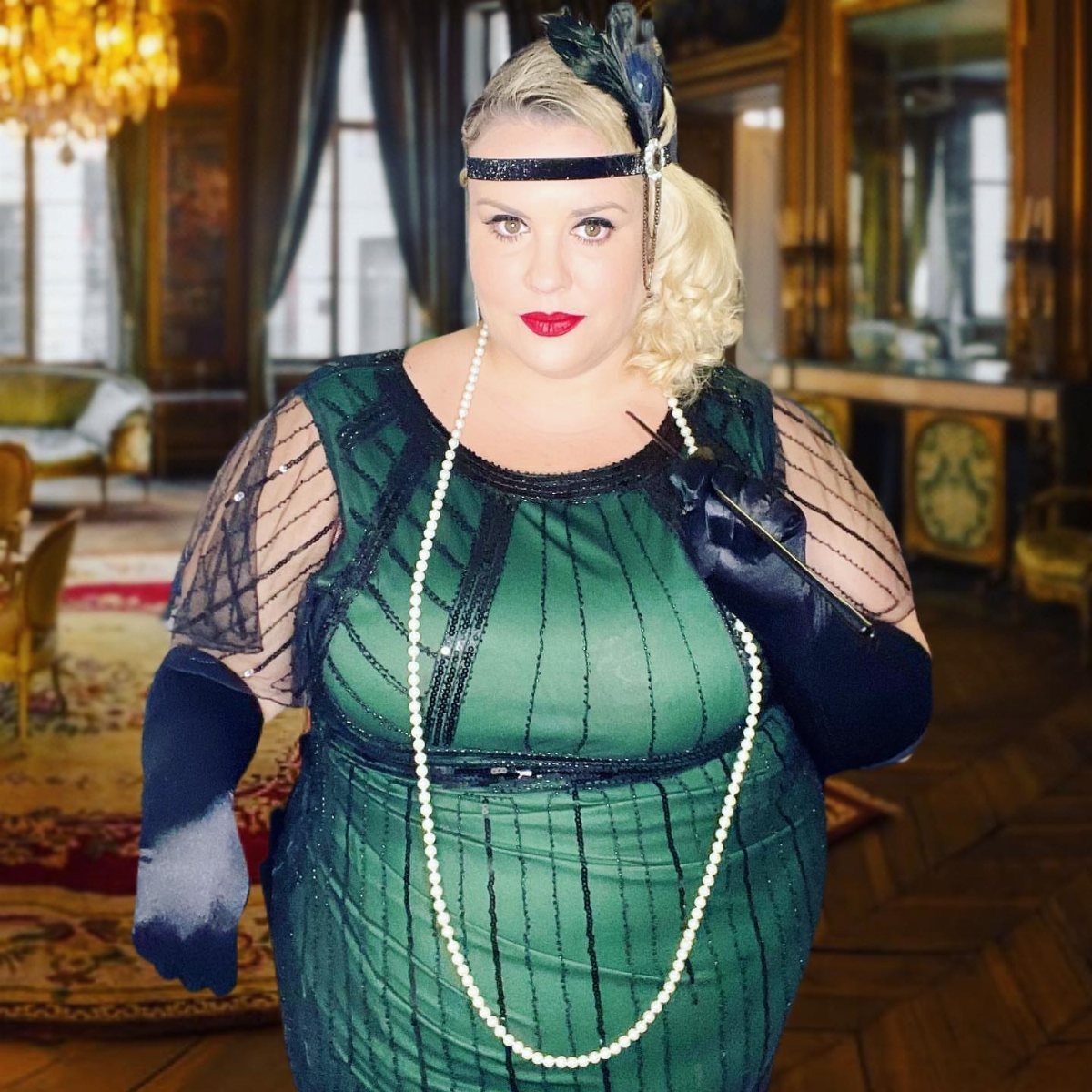 A woman in a green flapper dress, black gloves, and pearls posing indoors.