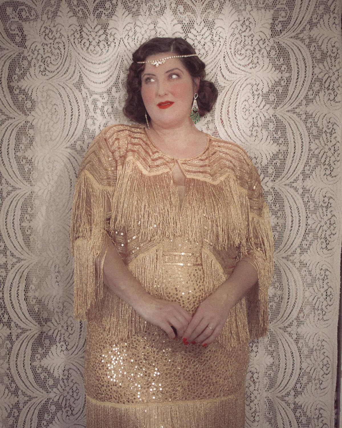 A woman in a gold sequined fringe flapper dress and headband posing against lace.