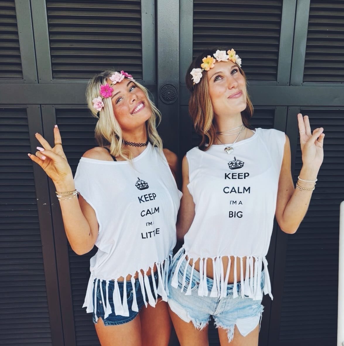 Two smiling women wearing flower crowns, white fringed tops, and denim shorts posing together.