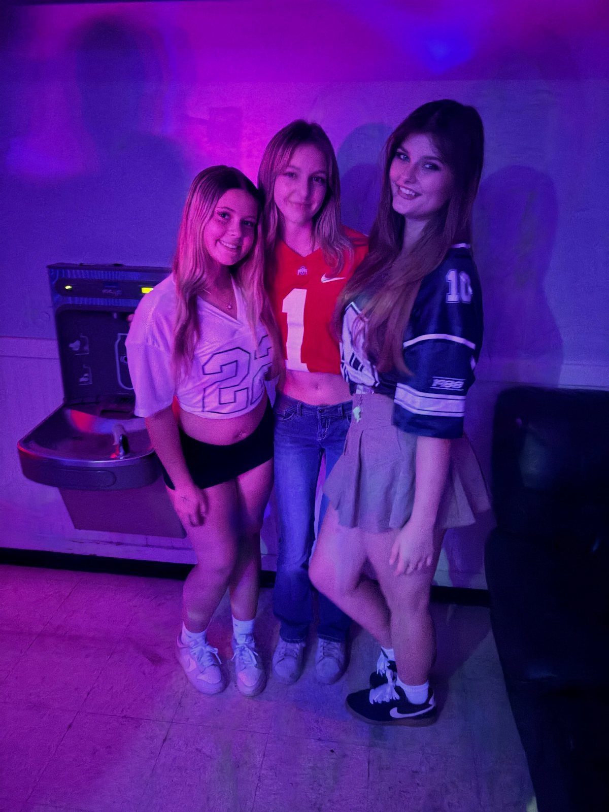 Three young women posing together in jerseys and casual clothing under purple light.