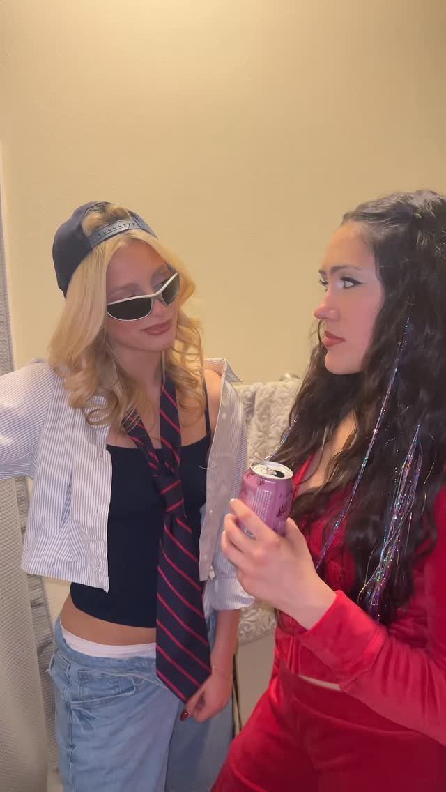Two women posing together, one wearing a striped tie, sunglasses, and an open button-down shirt.