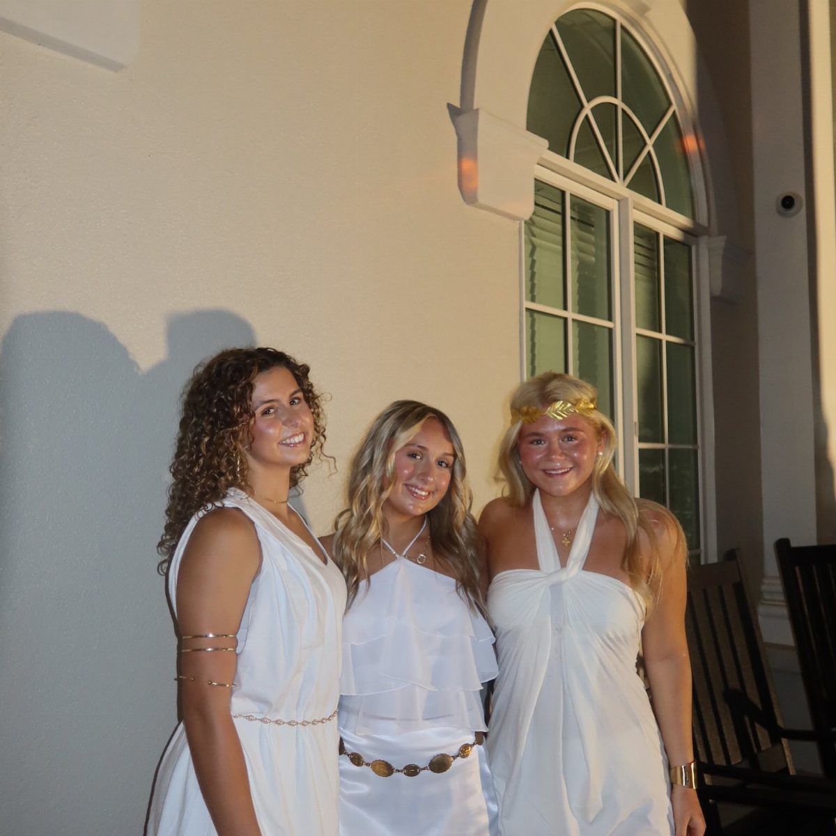 Three young women posing in white toga-style dresses and gold accessories at night.