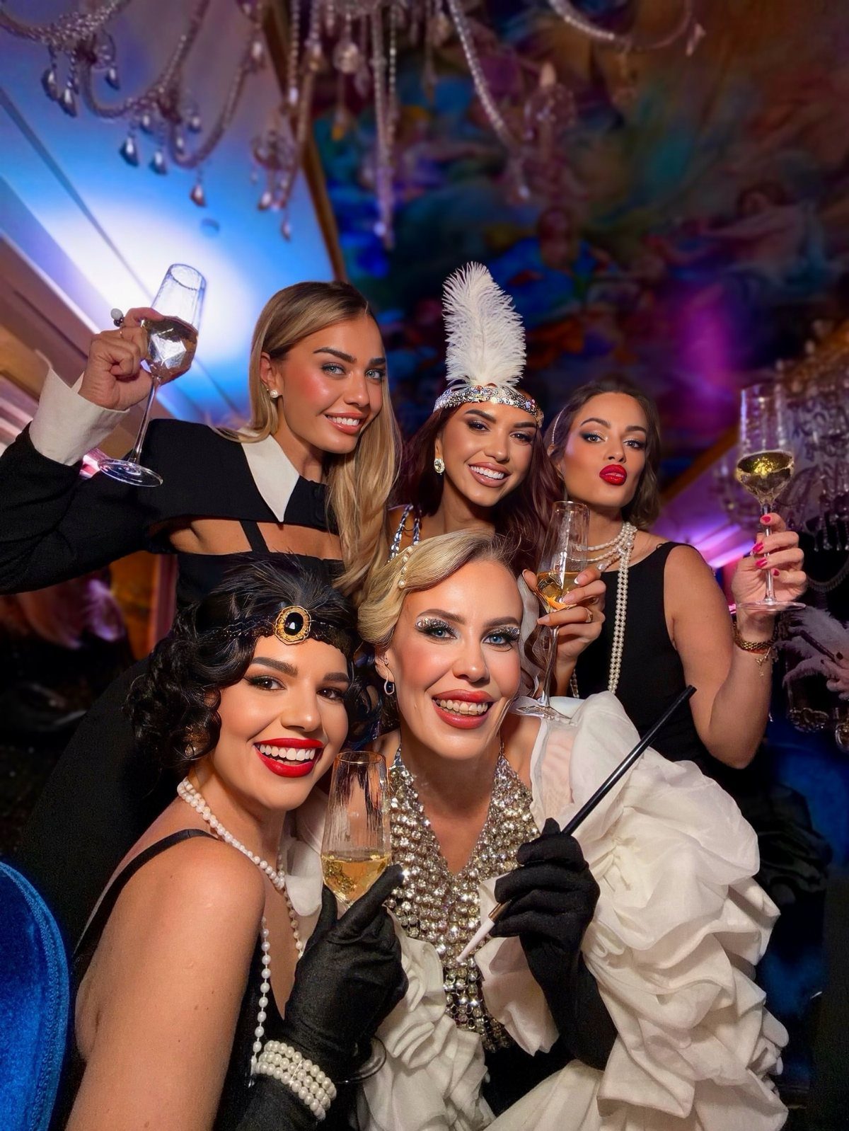 Five women dressed in vintage 1920s party attire holding champagne glasses under a chandelier.