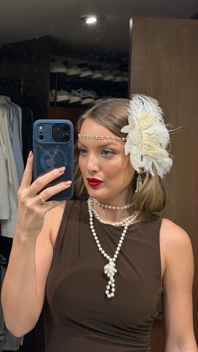 A woman with styled blonde hair wearing a large white feather accessory and pearl headband.