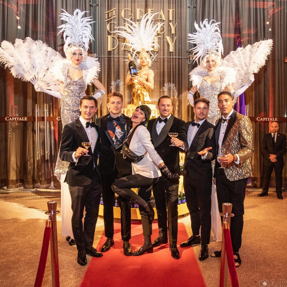 Men in black tuxedos and women in feathered costumes posing on a red carpet.