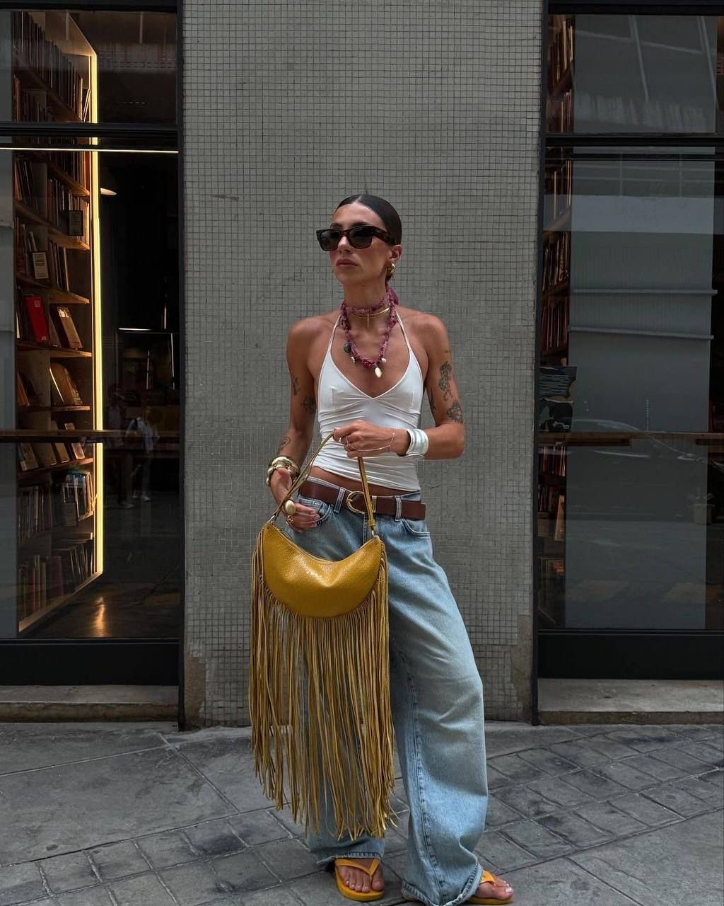 Woman in white tank top and wide-leg jeans carrying a large yellow fringed bag.