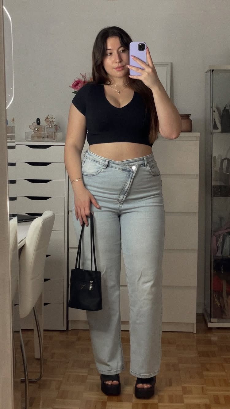 Woman in a black cropped top and high-waisted light blue jeans holding a black handbag.