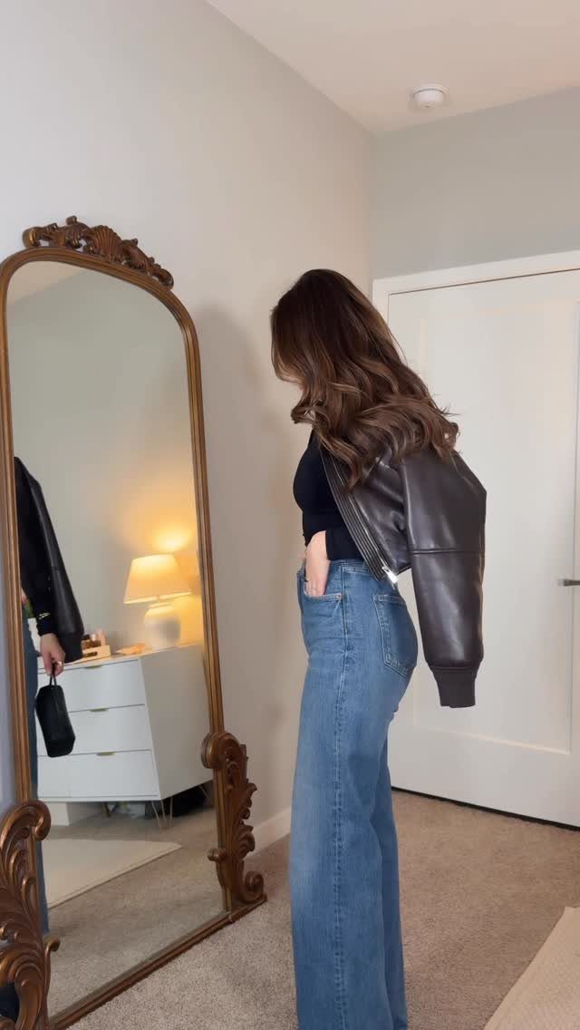 A woman wearing blue flared jeans and a black leather jacket standing near a large gold mirror.