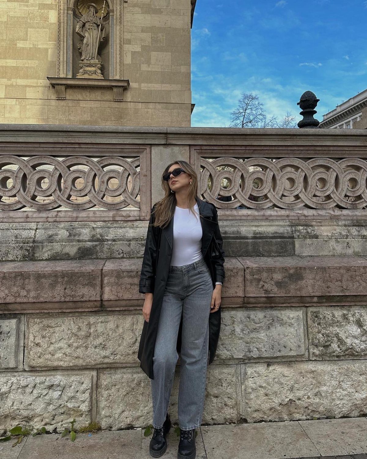 Woman in a black coat, white top, and grey jeans posing against a stone wall.