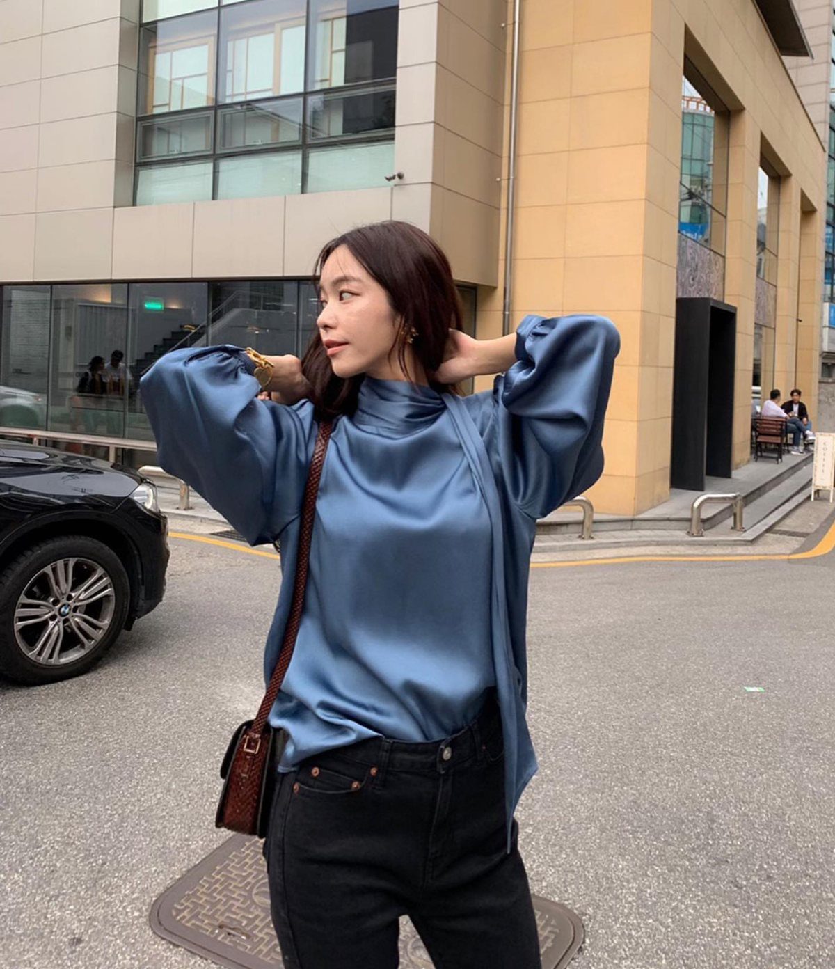 A woman wearing a blue satin blouse and black jeans posing on a city street.