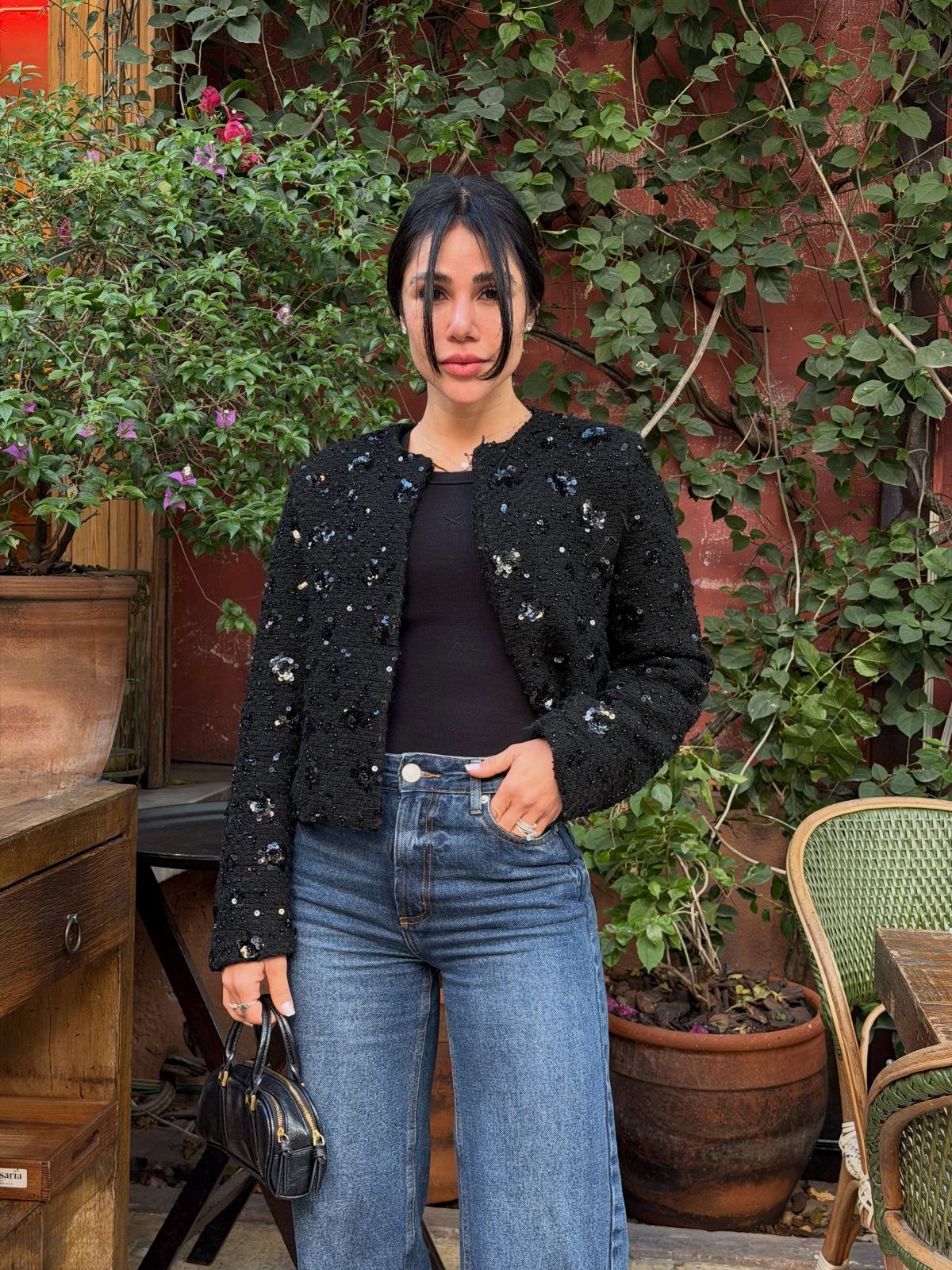 Woman wearing a black sequined jacket and blue wide-leg jeans standing near green plants.