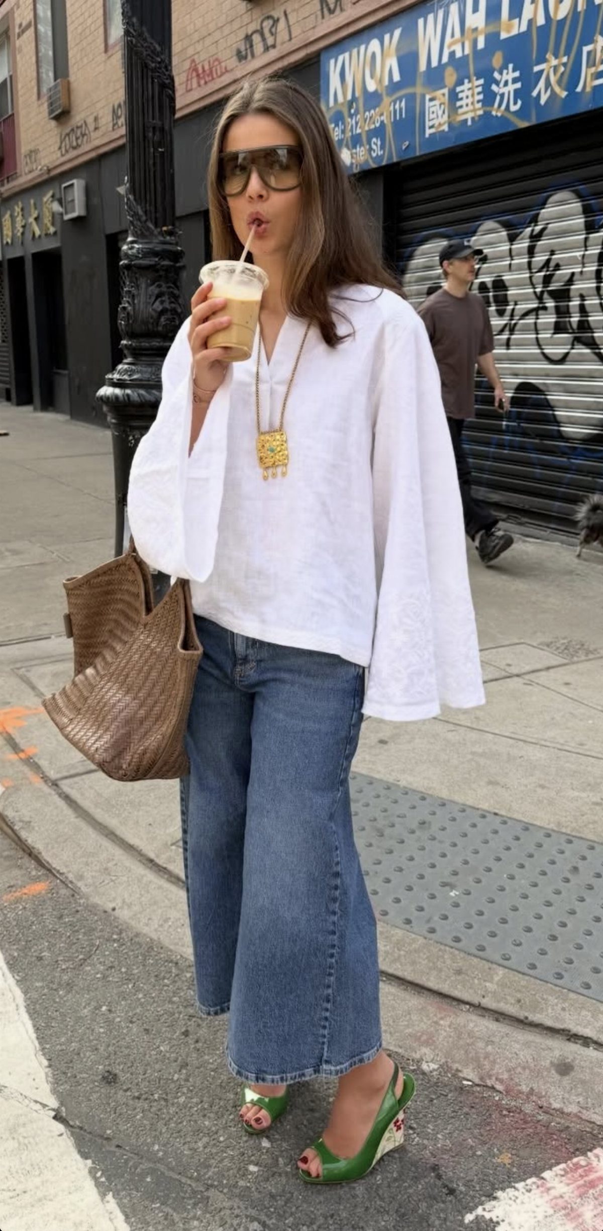 Woman in white blouse and wide-leg jeans standing on a city sidewalk holding a drink.