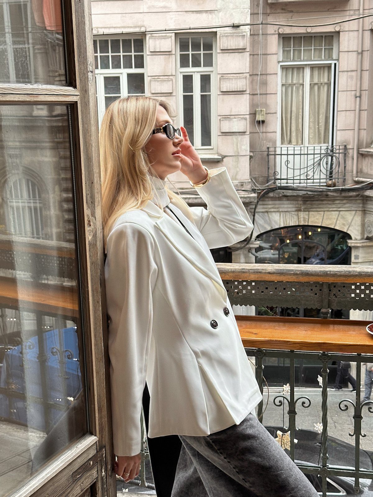 Blonde woman in a white tailored blazer and grey jeans leaning against a balcony window.