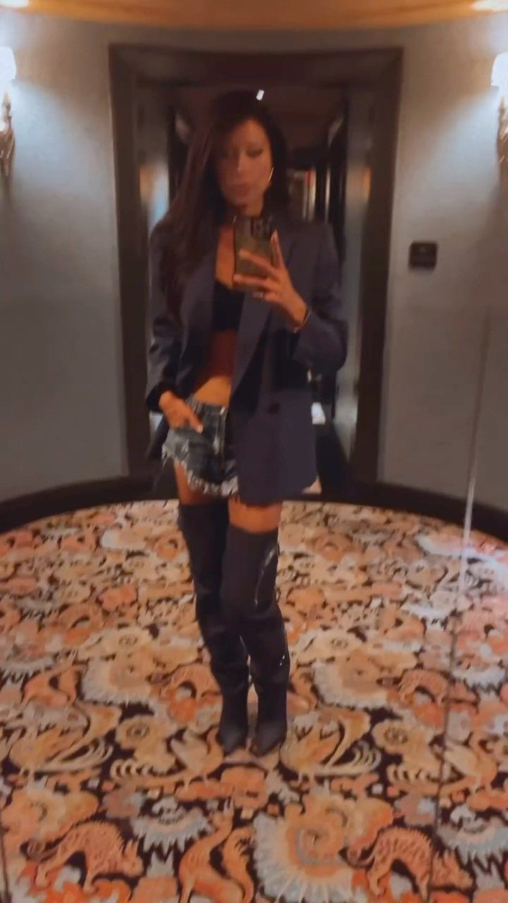 Woman in a blazer, denim shorts, and tall black boots taking a mirror selfie.