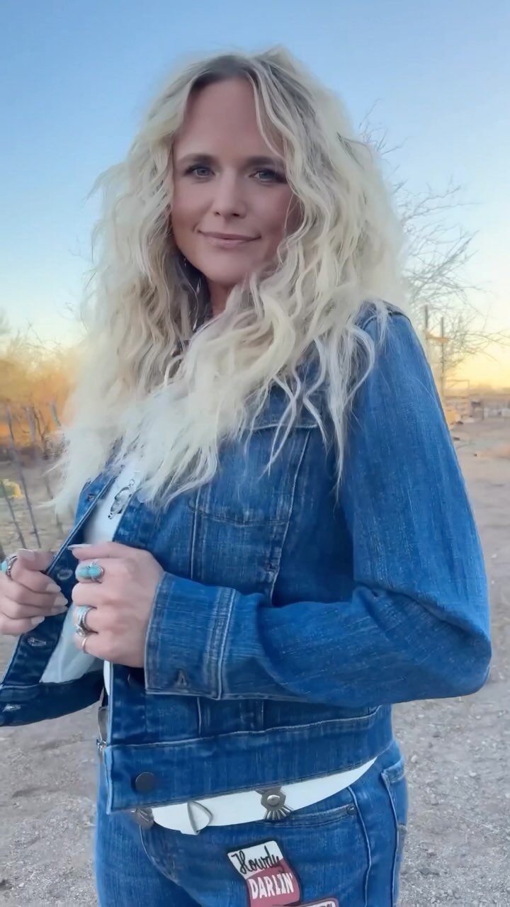 A blonde woman wearing a denim jacket, white graphic tee, and blue jeans outdoors.