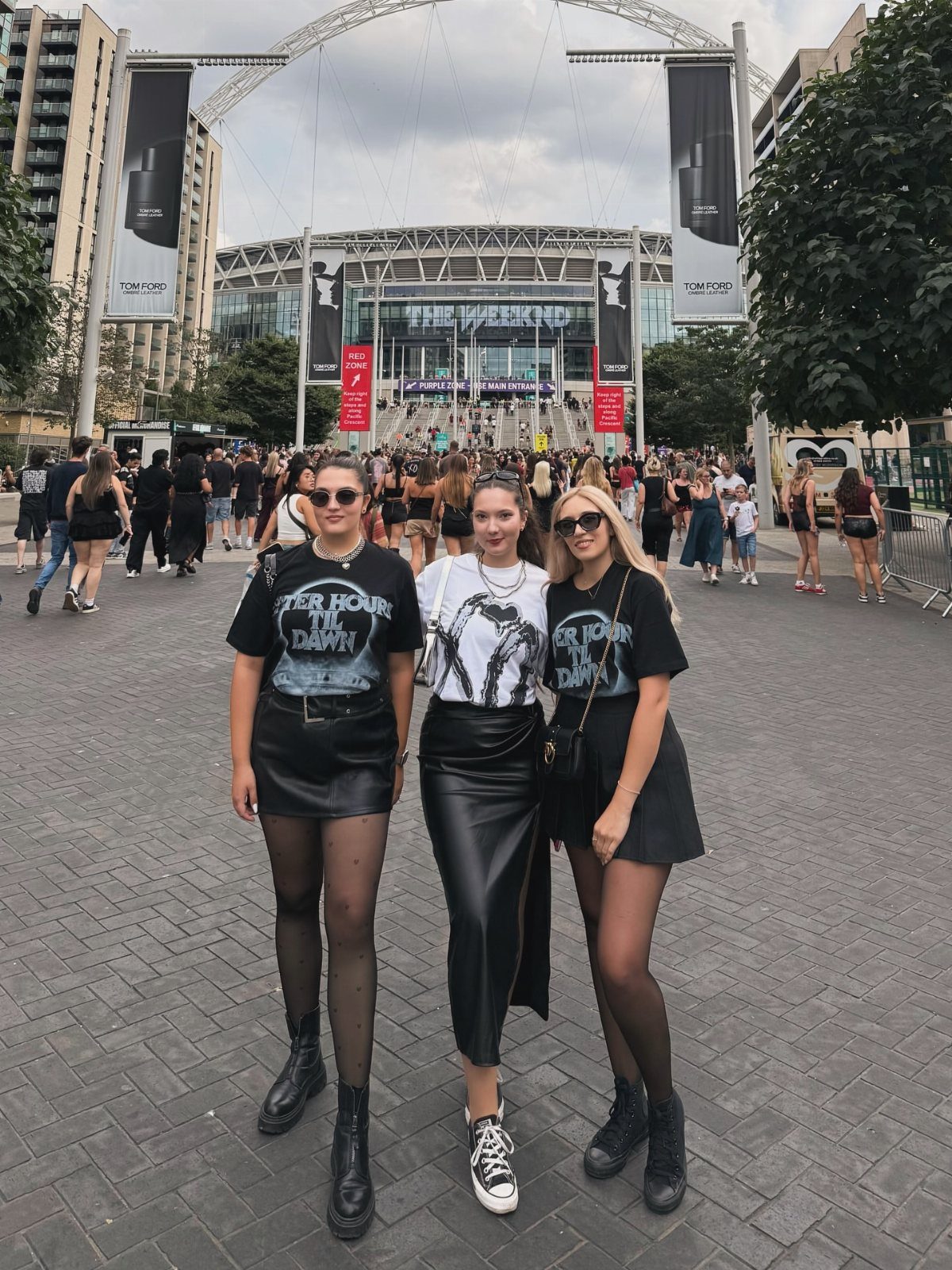 Three women posing in graphic tees, black skirts, and boots in front of a large stadium.