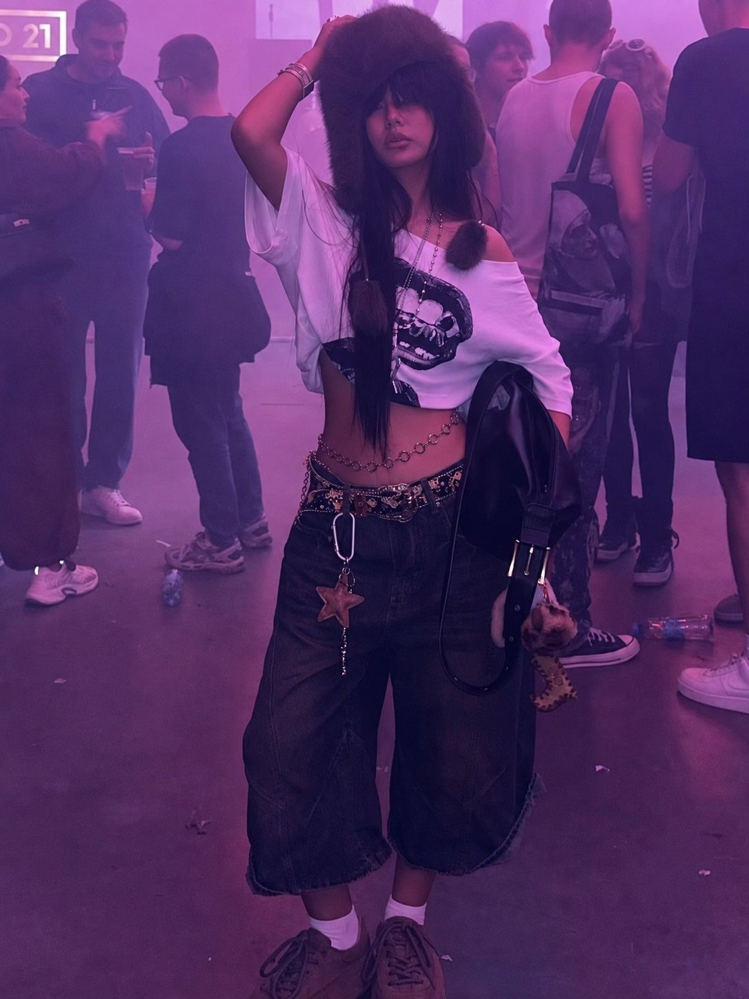 A woman in streetwear posing in a purple-lit concert venue wearing a graphic crop top and baggy shorts.