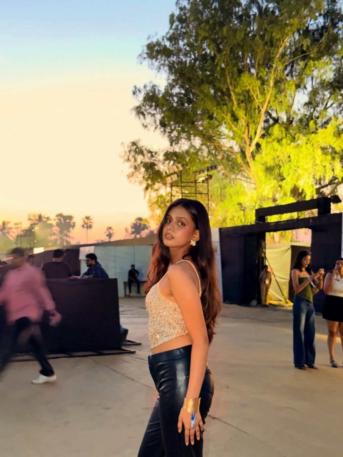 A woman in a sequined tank top and black pants posing outdoors at sunset.
