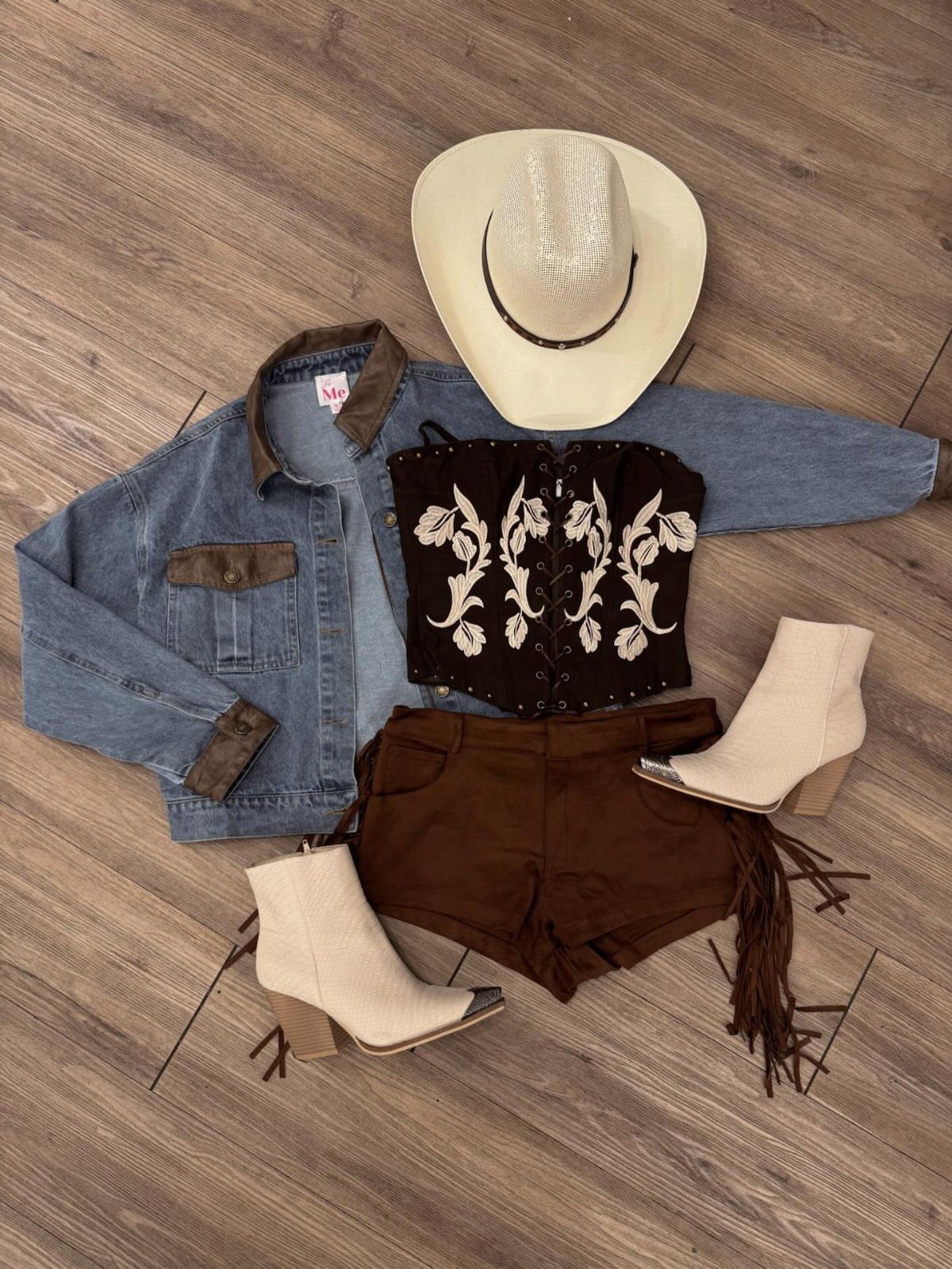 Flat lay of a denim jacket, embroidered corset, brown fringe shorts, cream boots, and cowboy hat.