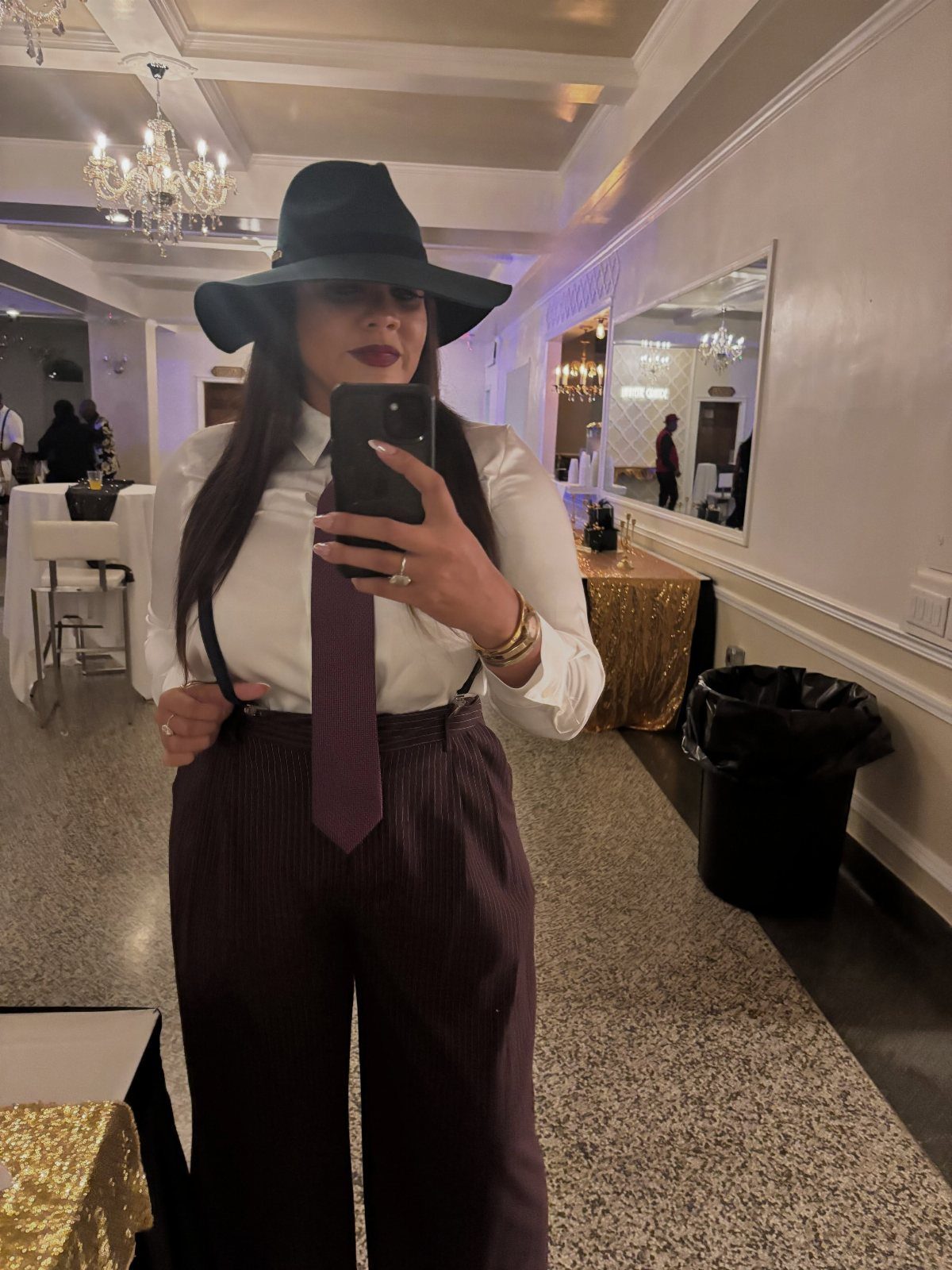 Woman in a white shirt, tie, suspenders, and wide-brimmed hat taking a mirror selfie.