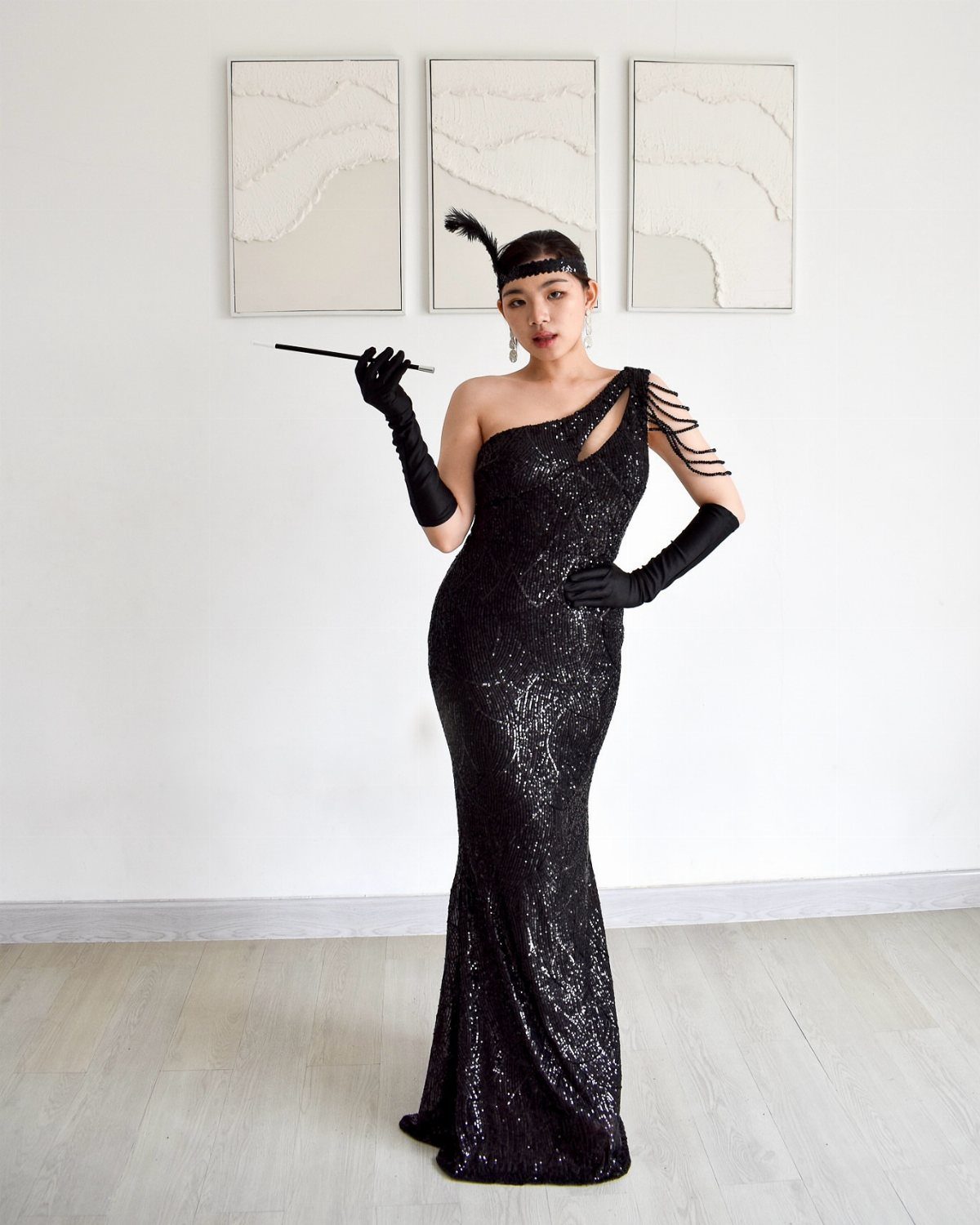 Woman in a black sequined one-shoulder gown, long gloves, and feathered headband posing against a white wall.