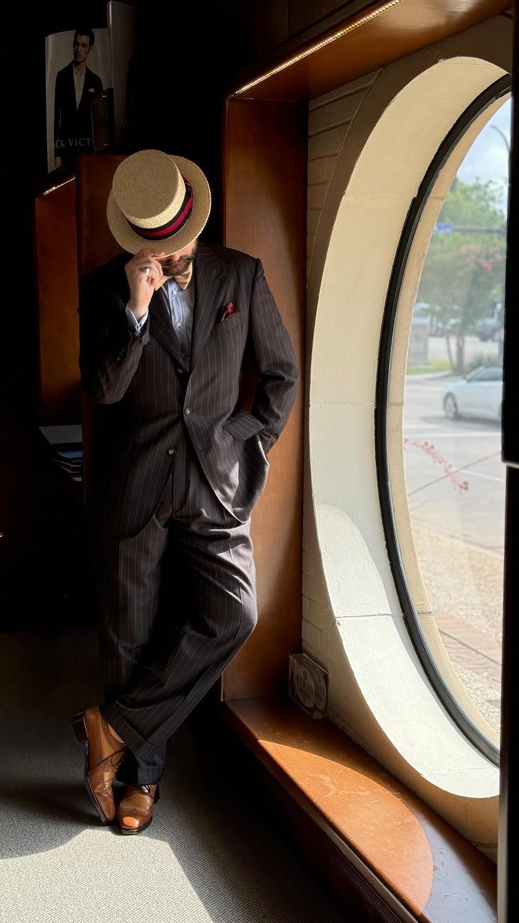 Man in a pinstripe suit and straw boater hat leaning against a large circular window.