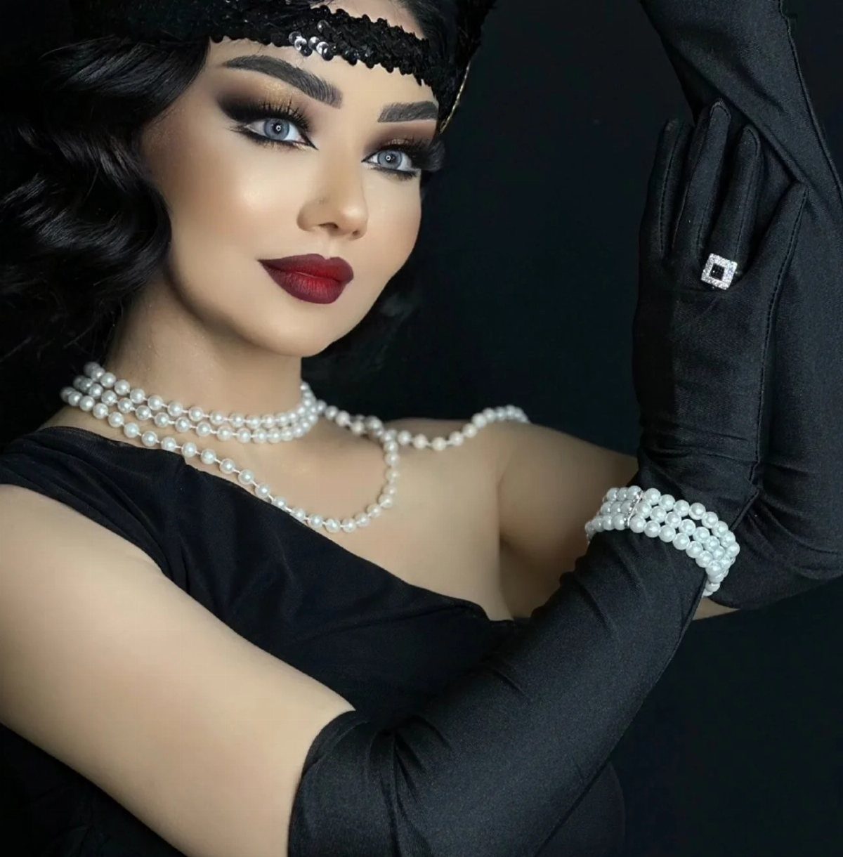 Woman wearing black satin long gloves, pearl necklaces, and a black headband against a dark background.
