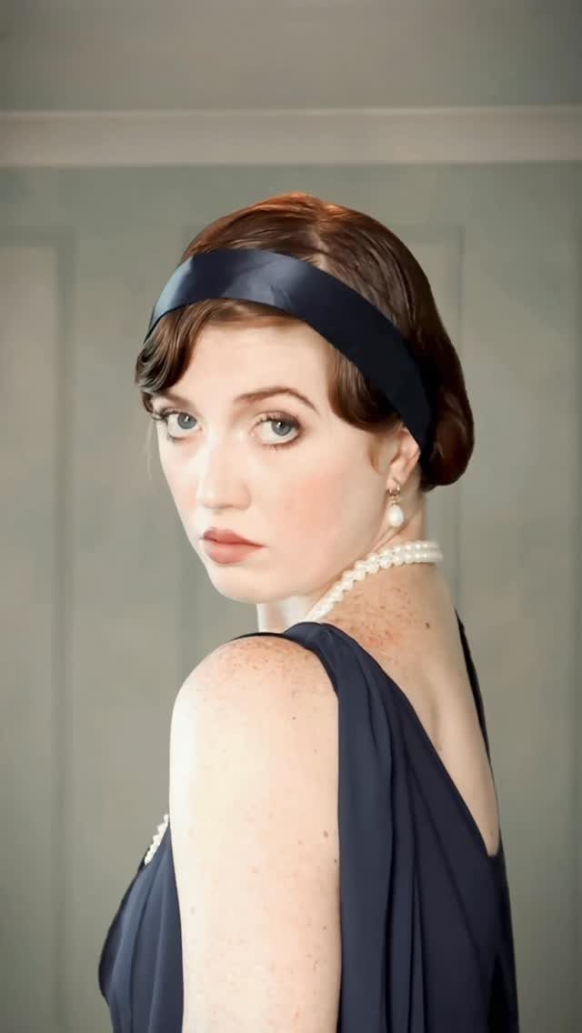 A woman in a 1920s style navy dress, headband, and pearls looking over her shoulder.