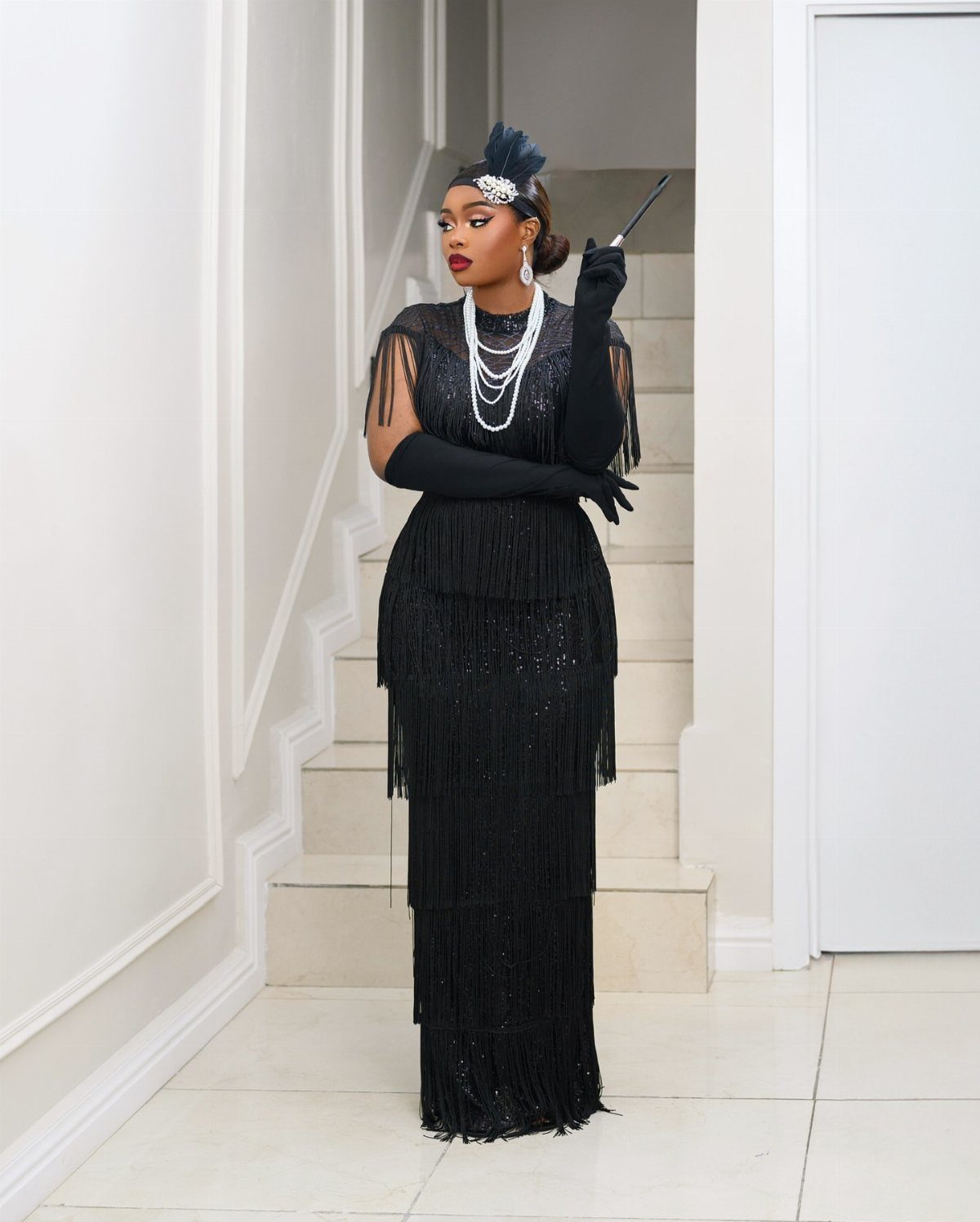 A woman in a black fringed flapper dress, long gloves, and pearls stands by a staircase.