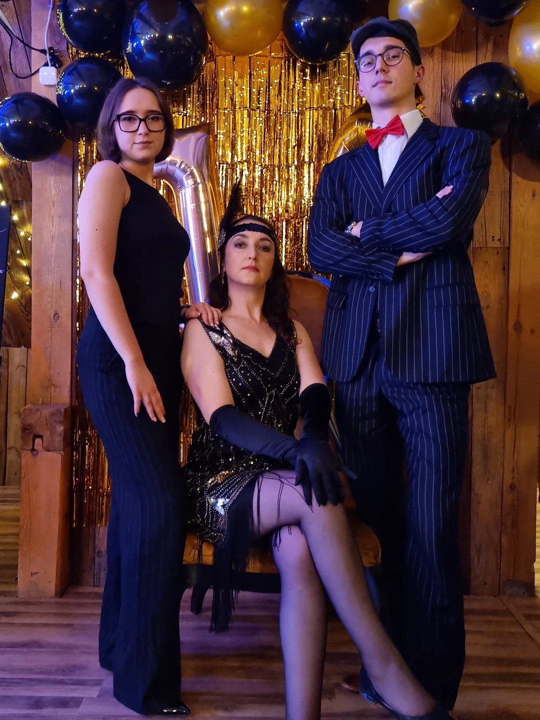 Three people dressed in 1920s flapper and vintage style attire posing against gold streamers.
