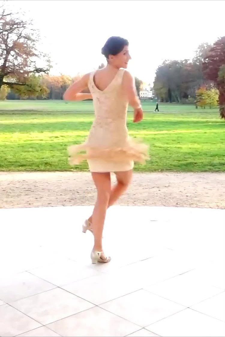 A woman in a beige fringed dress dancing on a white tiled patio in a park.
