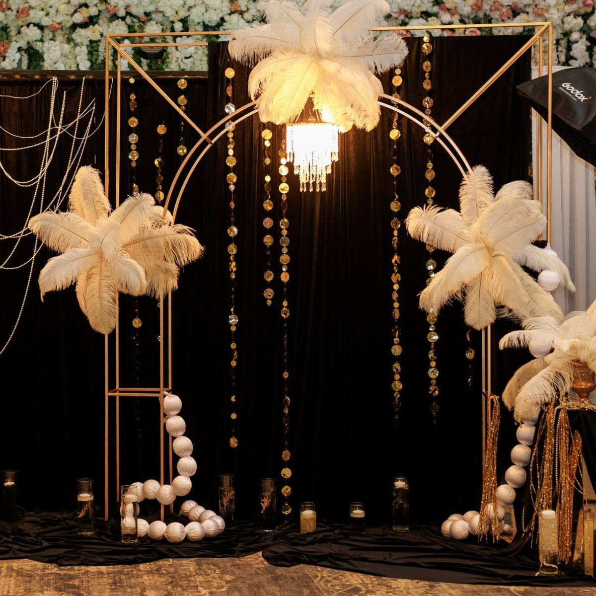 Gold archway decorated with white ostrich feathers, gold sequins, and a crystal chandelier against black fabric.