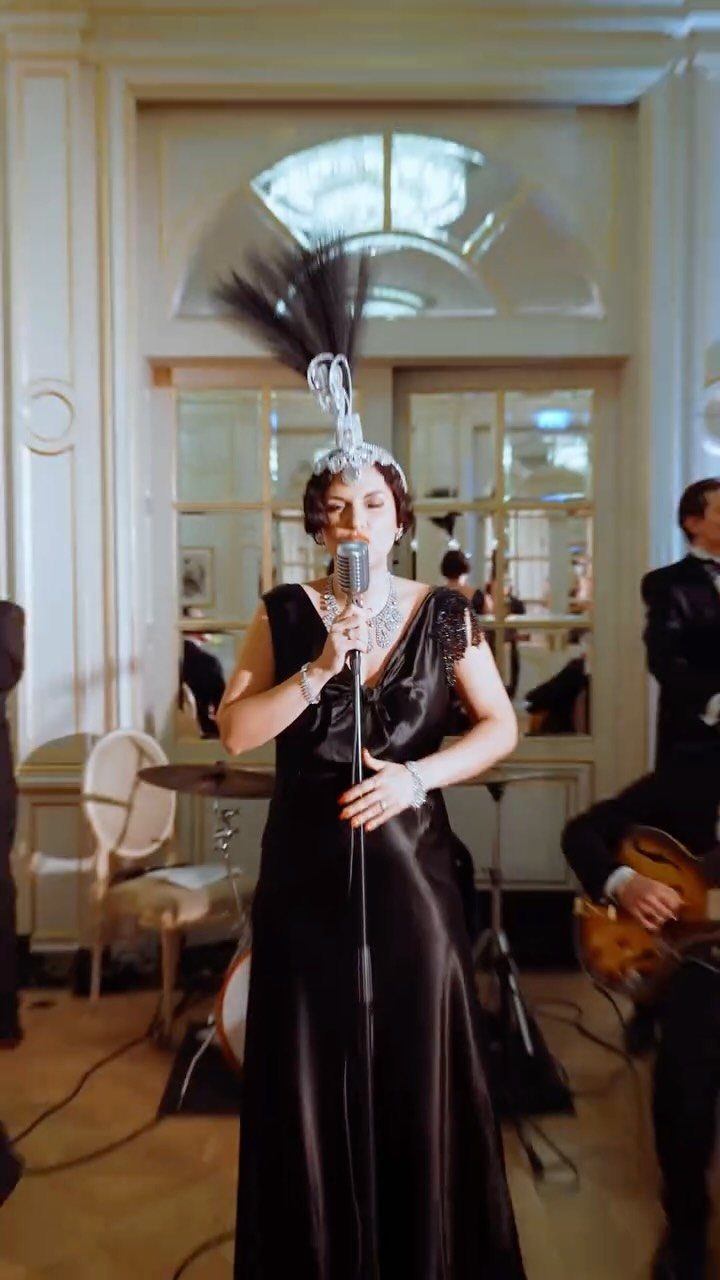 Woman in a black satin flapper gown singing into a vintage microphone in an elegant room.