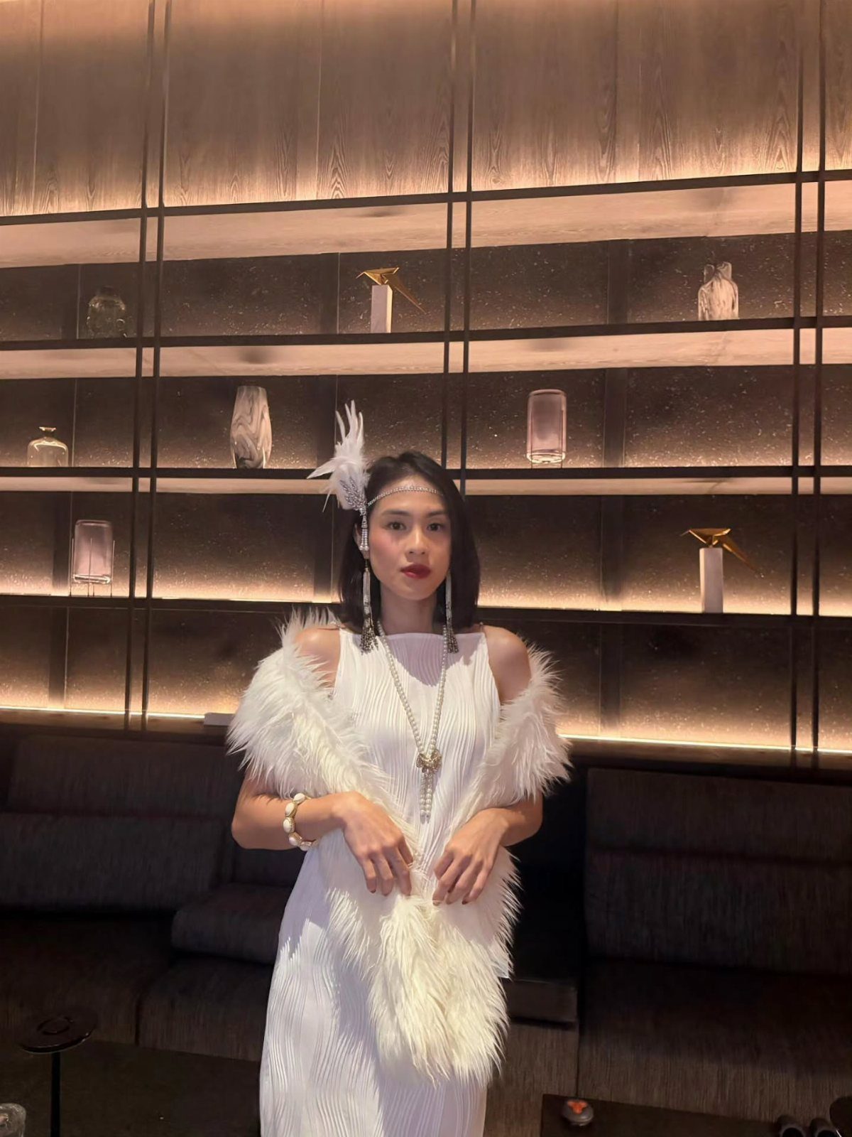 A woman in a white feathered flapper gown and pearl necklace posing against a backlit shelf.