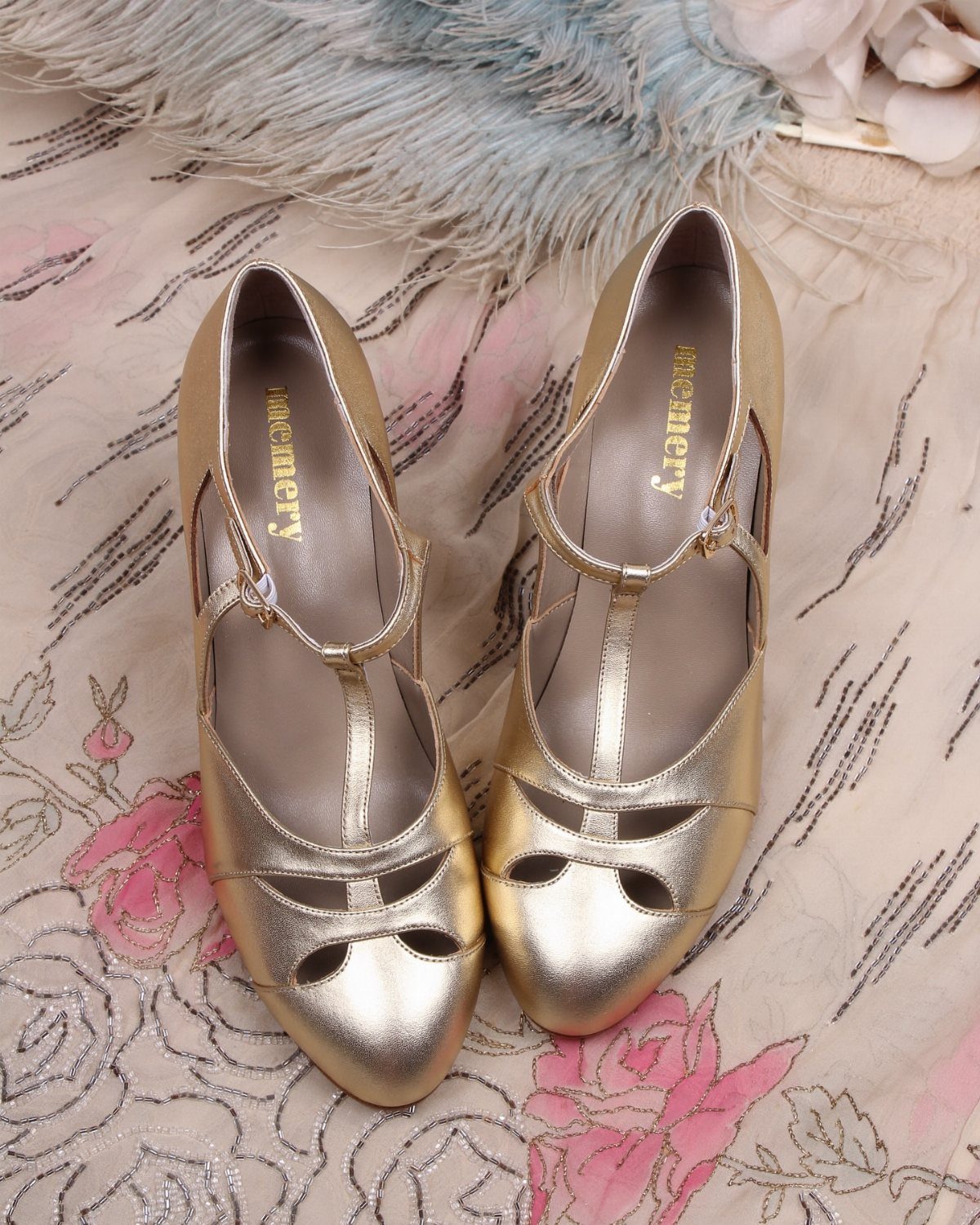 Gold metallic T-strap flapper shoes resting on floral embroidered fabric with blue feathers.
