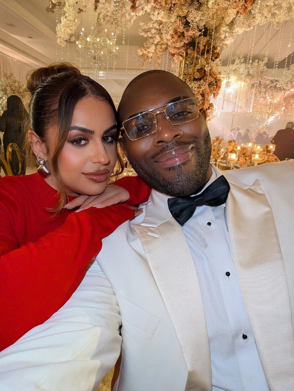 A man in a white tuxedo and a woman in red posing in a decorated ballroom.