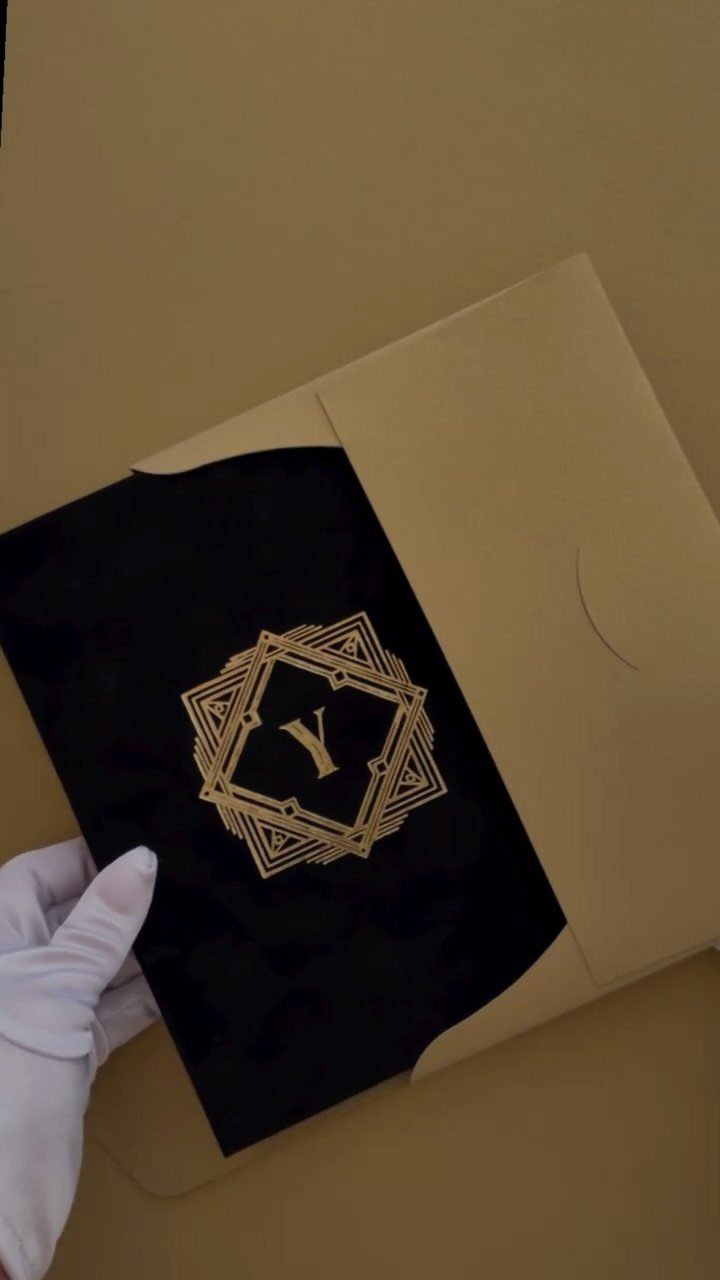 A gloved hand pulls a black Art Deco themed invitation with gold lettering from a gold envelope.