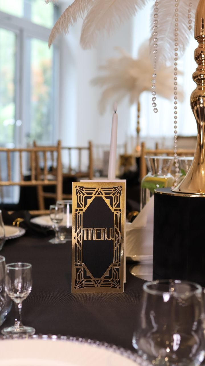 Art Deco style menu card on a black tablecloth with feathers and glassware.