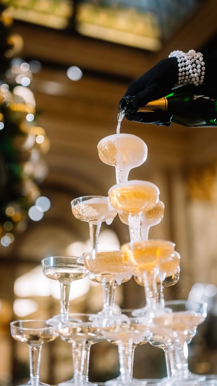 A gloved hand pours champagne into a pyramid of sparkling coupe glasses forming a tower.