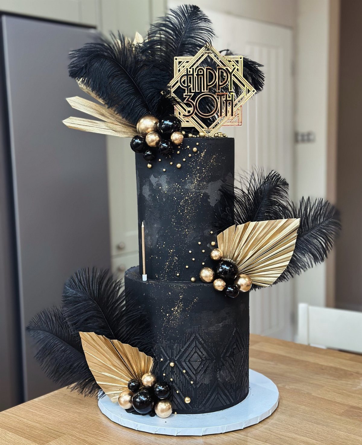 Two-tier black cake with gold speckles, black feathers, and gold geometric decorations on a wooden table.