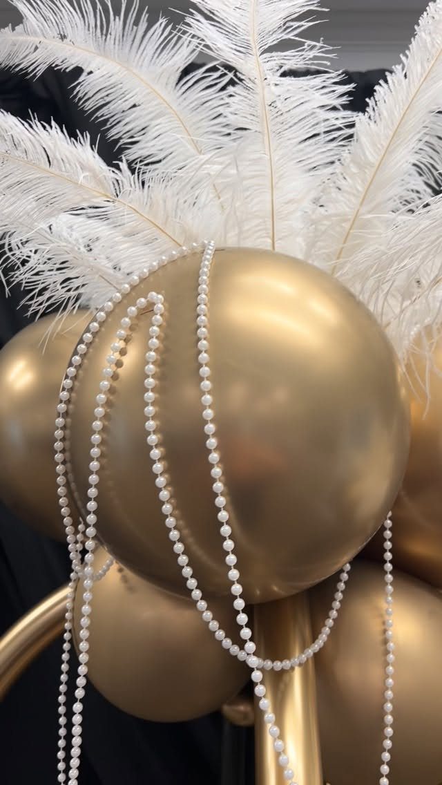 Gold balloons adorned with white ostrich feathers and white pearl bead strands.