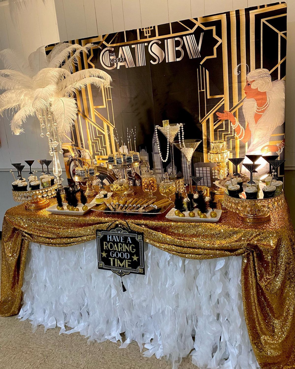 A decorated dessert table with gold sequins, white fringe, and ostrich feathers for a Gatsby party.