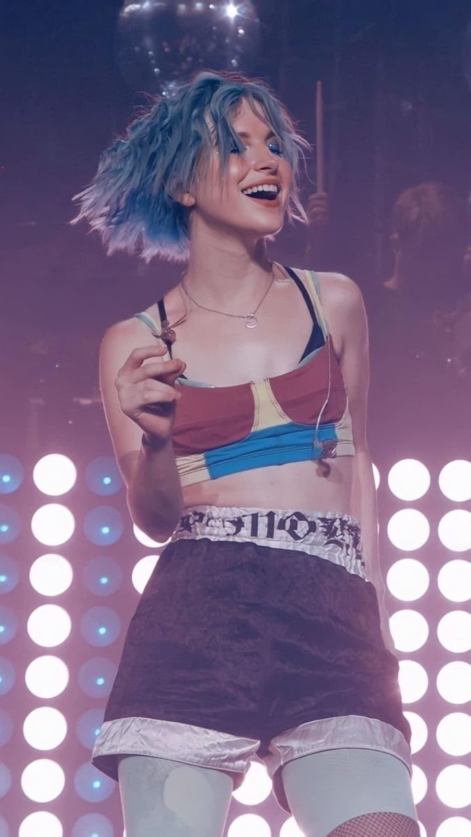 A woman with blue hair smiling while wearing a color-blocked cropped tank top on stage.