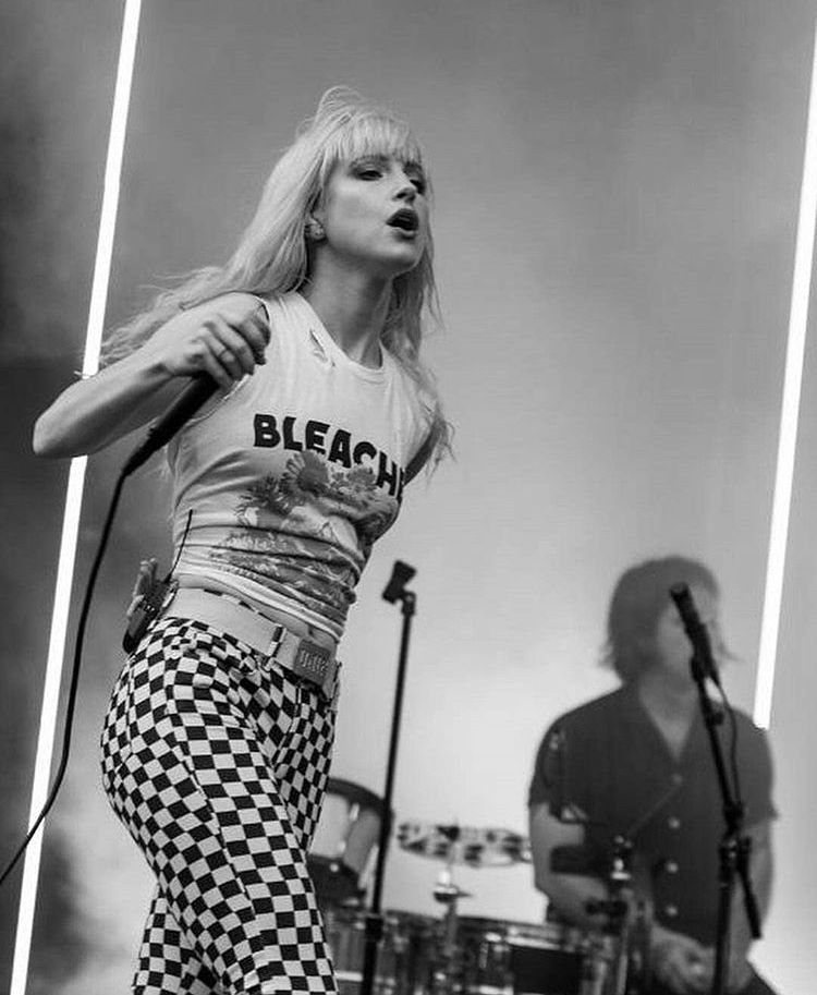 A blonde woman singing into a microphone while wearing a white graphic tank top.