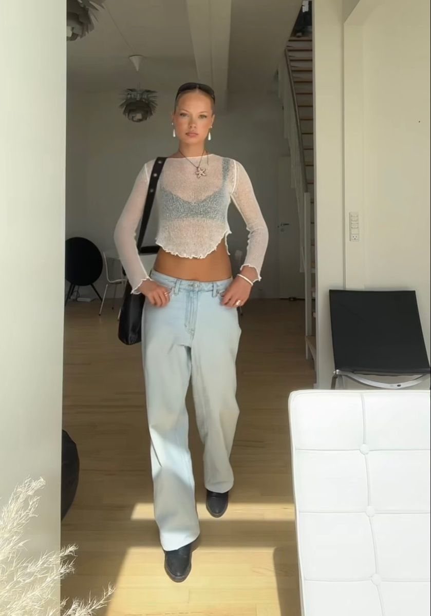 Woman wearing a sheer white mesh long-sleeve crop top and light blue wide-leg jeans walking indoors.