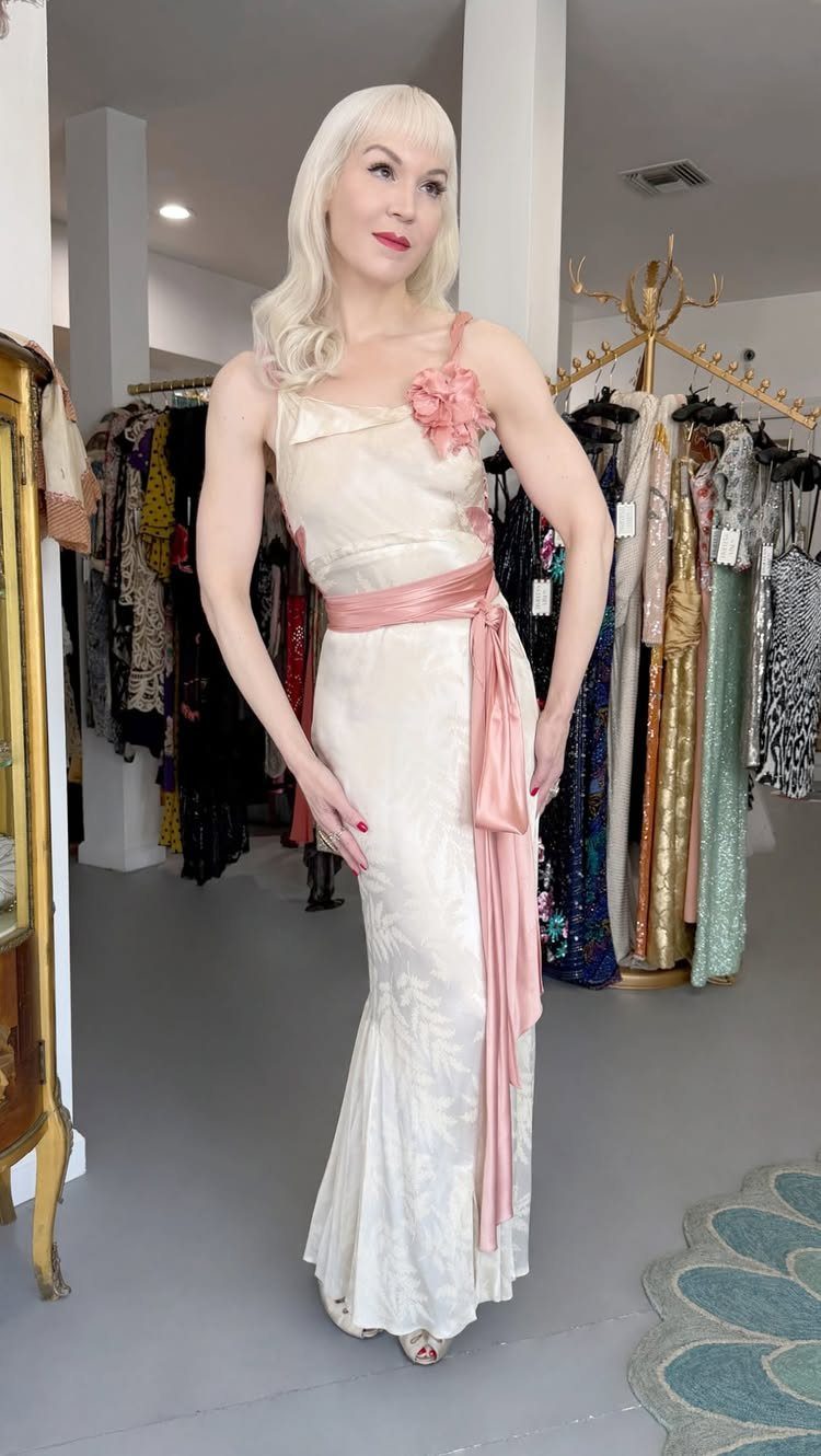 Blonde woman posing in a cream-colored bias-cut gown with a pink satin sash.