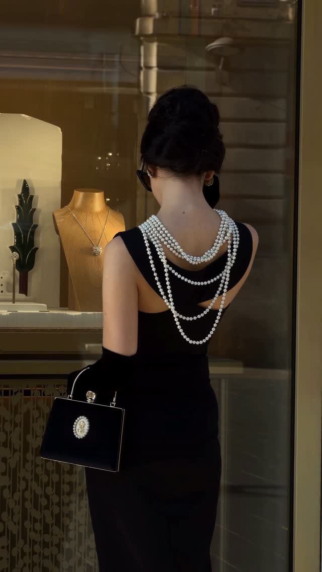 Woman in a black dress and pearls holding a handbag while looking into a window.