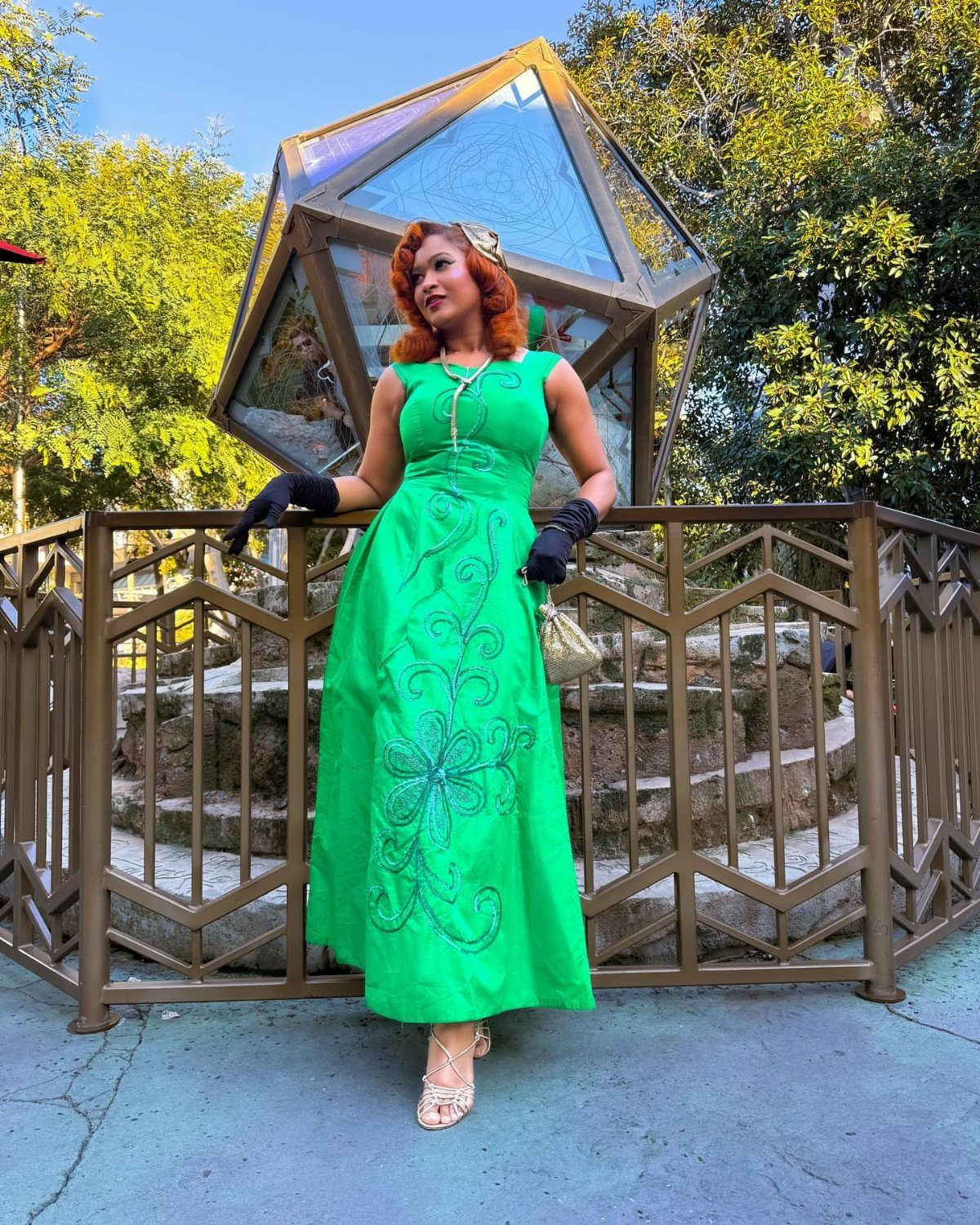 A woman wearing a vintage green gown and black gloves posing outdoors near a glass structure.