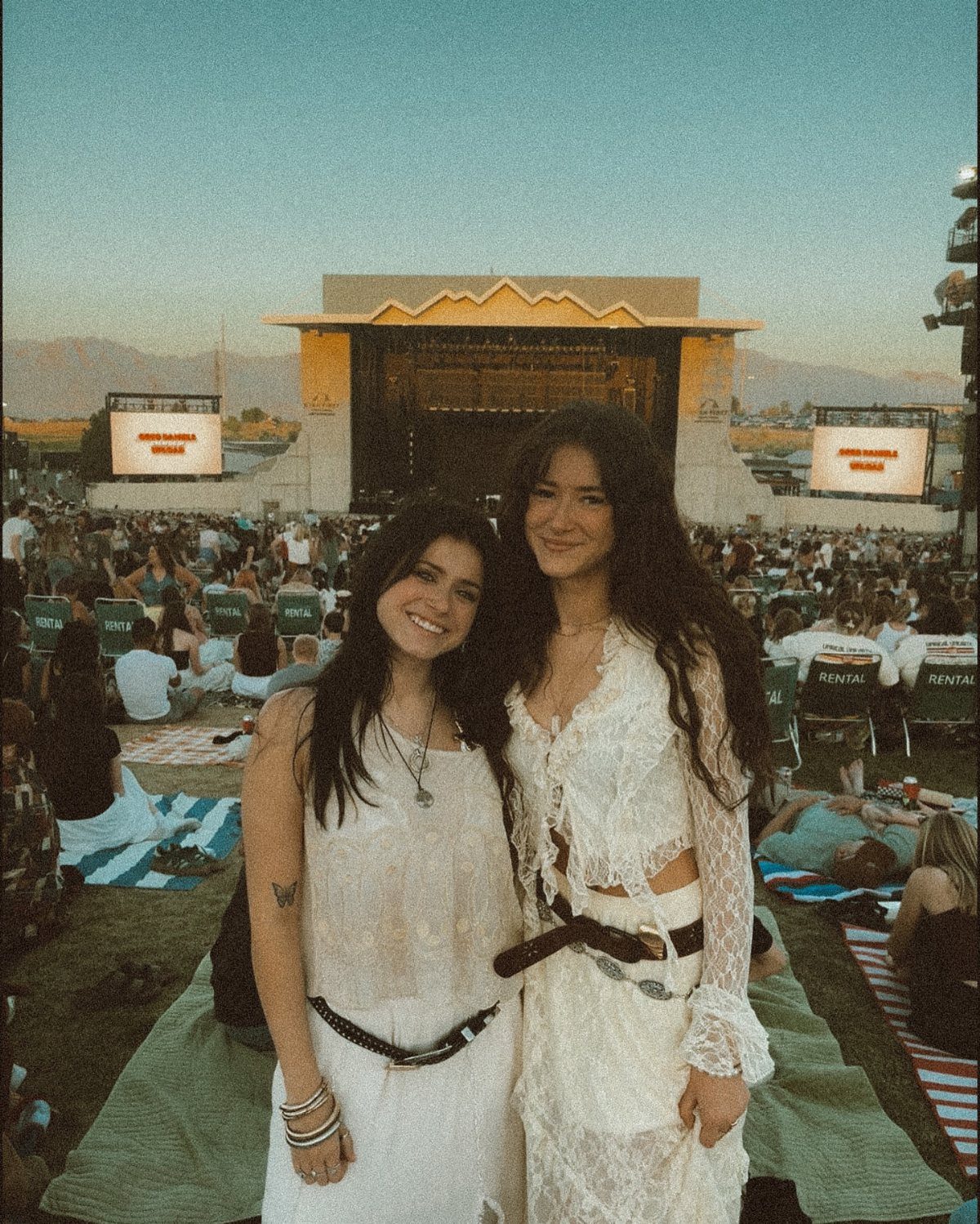 Two women in white bohemian outfits posing at an outdoor music festival during the day.