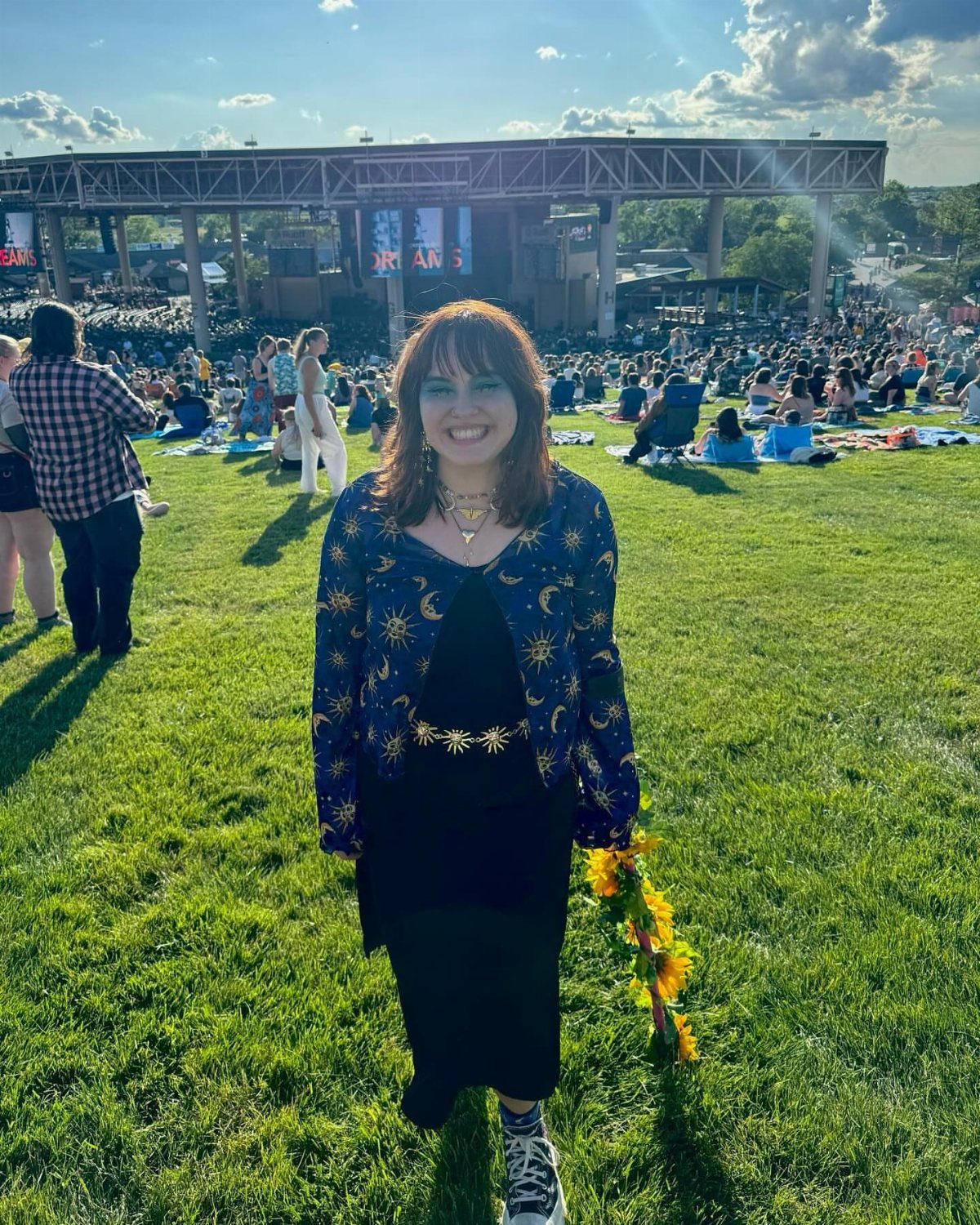 A woman in a celestial patterned cardigan and black dress smiling in a grassy concert field.